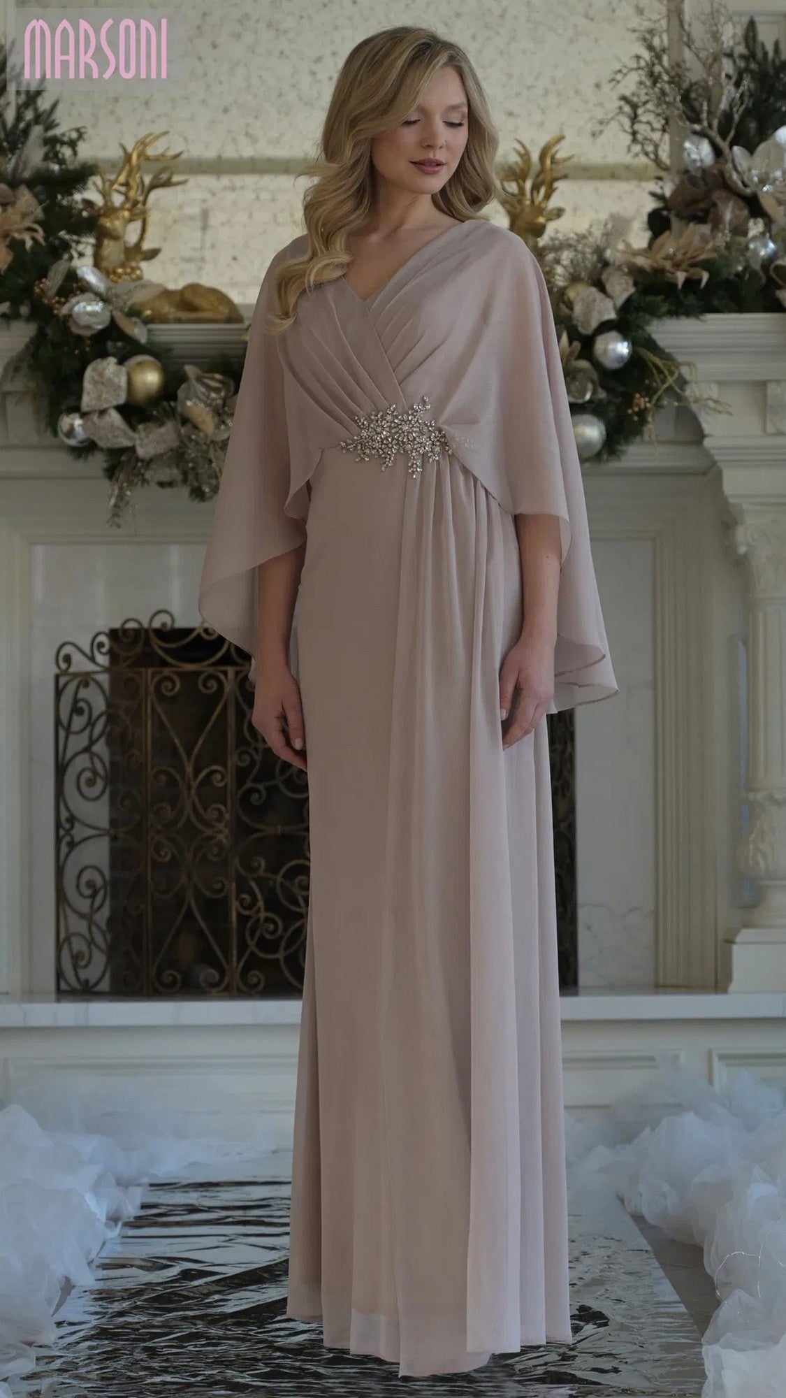 A woman poses gracefully indoors before a decorated fireplace, wearing the Marsoni MV1361 Long Formal Dress in champagne, featuring cape sleeves and a crystal-beaded applique at the waist.